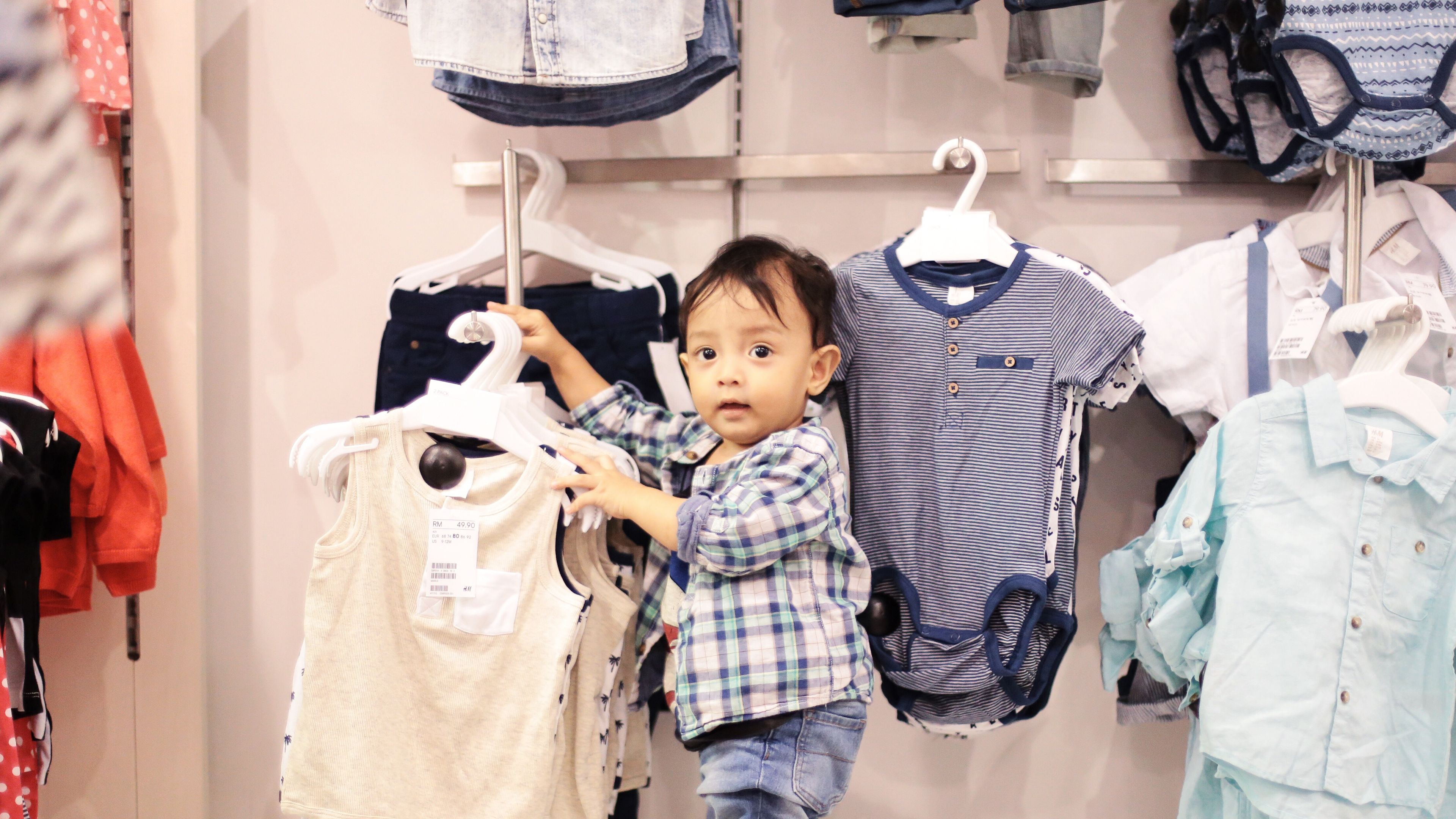 A toddler in a plaid shirt shopping for clothes, holding a hanger and looking curiously at the camera.